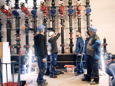 Mehrere Handwerker von Brasst stehen vor großen Rohrleitungen in einem Raum und beratschlagen sich.